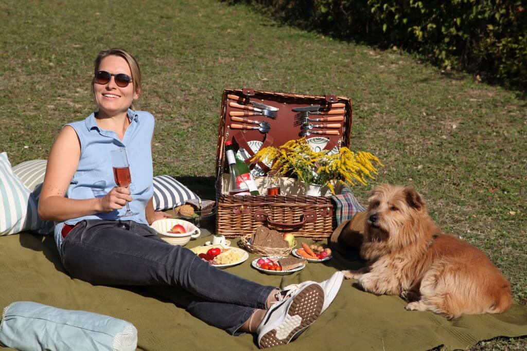 Weinviertler Picknick mit Hund. Frau mit Hund liegen auf der Picknickdeck mit gefüllten Picknickkorb und gefüllten Tellern.